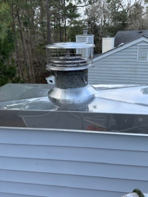A shiny stainless steel chimney cap with a protective wire mesh screen sits atop a metal chase cover on a house roof - https://www.juniatachimneyinspectionandrepair.com/