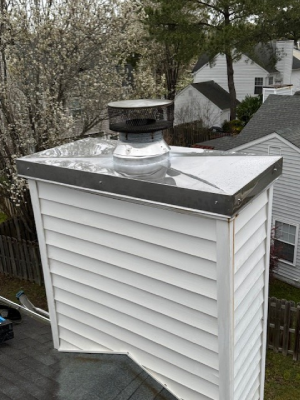 A chimney with white horizontal siding is topped with a shiny metal chase cover and a black mesh spark arrestor - https://www.juniatachimneyinspectionandrepair.com/