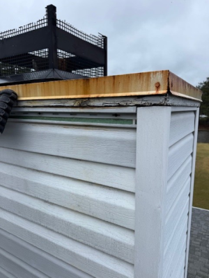 A close-up of a chimney structure with white vinyl siding, showing significant rust on the metal cap and deterioration of the wood trim directly underneath it - https://www.juniatachimneyinspectionandrepair.com/