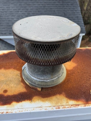 A weathered metal chimney cap with a wire mesh screen sits atop a heavily rusted metal chimney crown - https://www.juniatachimneyinspectionandrepair.com/